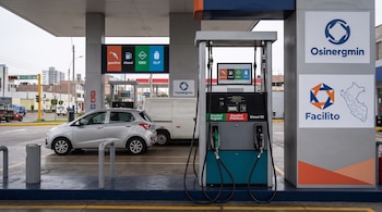 Surtidor de combustible en primer plano con logotipos de Osinergmin y Facilito, pantallas informativas y dos automóviles estacionados al fondo.