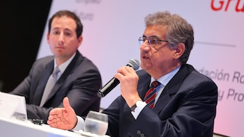 Un hombre con anteojos y traje habla por un micrófono sentado a una mesa de conferencia junto a otro asistente