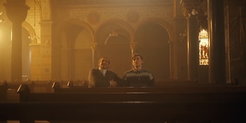 Dos hombres se encuentran sentados en una banca de madera dentro de una iglesia con arcos, columnas e iluminación cálida