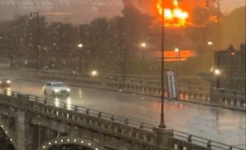 Puente de carretera con coches y farolas encendidas bajo la lluvia en una zona metropolitana, con un gran resplandor naranja de fuego en el fondo