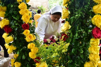 Primer plano de una monja en hábito blanco ajustando rosas rojas, amarillas, rosadas y blancas en un arreglo floral, con columnas de rosas a los lados