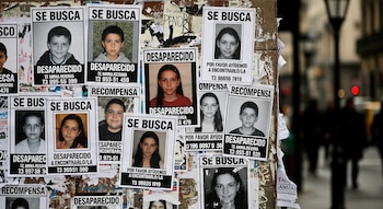 Primer plano de un muro o poste cubierto con múltiples carteles de "Se Busca" que muestran fotos de niños y adolescentes desaparecidos, con una calle borrosa al fondo.