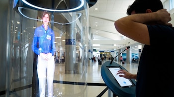 Persona de perfil opera pantalla táctil frente a tubo transparente con holograma de figura humana en uniforme azul dentro de terminal de aeropuerto.