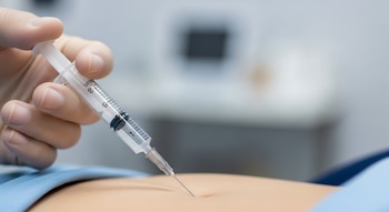 Una mano con guante blanco sostiene una jeringa con una aguja insertada en el abdomen de una persona.