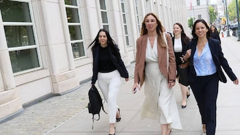 Cuatro mujeres caminan en una acera de adoquines, vistiendo ropa formal de negocios frente a un edificio claro con ventanas rectangulares