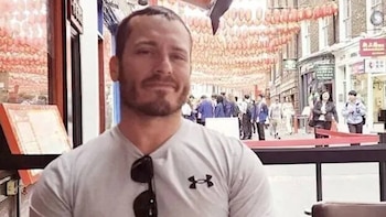 Hombre de tez blanca, barba oscura y camiseta gris con el logo de Under Armour sonríe levemente; de fondo, una calle urbana con farolillos rojos y personas