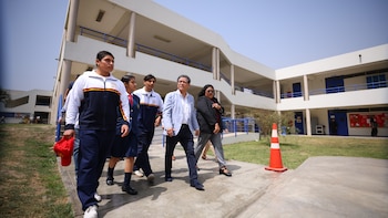 José Antonio Chang, Rebeca Ata Muñoz y estudiantes caminan por un pasillo de concreto. Hay edificios blancos con barandas azules, césped y un cono