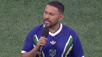 Hombre de barba corta y arete, viste camiseta deportiva azul con logos Adidas y MLS, sujeta un micrófono negro. Fondo de césped verde.