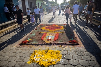 Un hombre con gorra azul crea una alfombra de aserrín colorida en el suelo, con diseños de un sol radiante y dos estructuras arquitectónicas coloniales, en una calle empedrada