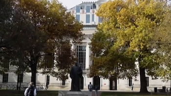 Estatua en plaza frente a un edificio de universidad blanco con columnas, rodeado de árboles con hojas de otoño; varias personas caminan por el campus