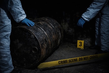 Un forense con guante azul toca un barril metálico oscuro y oxidado. Una cinta amarilla de "CRIME SCENE" y un marcador de evidencia "3" son visibles en el suelo oscuro.
