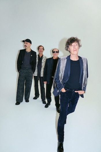 Los cuatro miembros de Cuarteto de Nos posan de pie en fila contra un fondo blanco. Visten trajes oscuros y elegantes, con detalles brillantes en uno