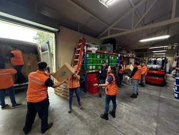 Varias personas con chalecos naranjas de SE-CONRED cargan cajas en una furgoneta blanca dentro de un almacén. Hay estanterías, un montacargas y palets