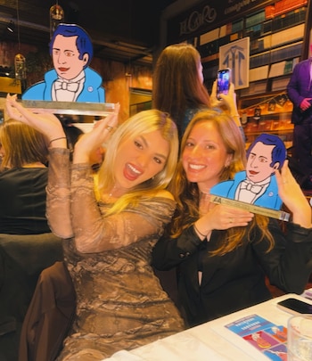 Dos mujeres sonrientes sostienen trofeos acrílicos con la ilustración de una figura histórica en una mesa de restaurante