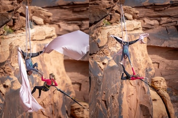 Dos acróbatas cuelgan de sedas y cuerdas, una invertida y la otra sosteniendo sus manos, frente a un imponente acantilado de roca arenisca en Utah