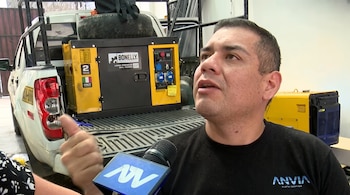 Un hombre habla frente a un micrófono junto a la parte posterior de una camioneta blanca que contiene un generador eléctrico.