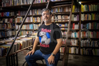 Un hombre con gafas, camiseta oscura con capibara y pantalón vaquero se sienta en una escalera de madera dentro de una librería con estantes repletos de libros