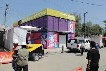 Un edificio colorido de contenedores con letrero 'Biciestacionamiento Universidad' bajo un cielo azul, con trabajadores y vehículos en una zona de obras