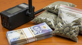 Un teléfono satelital de color negro, tres bolsas transparentes con marihuana y dos fajos de billetes de dos mil colones costarricenses sobre una mesa de madera.