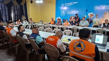 Un grupo de personas sentadas en mesas de un salón con chalecos naranja y azul. Al fondo, un banner azul de la Gobernación de Suchitepéquez, Guatemala