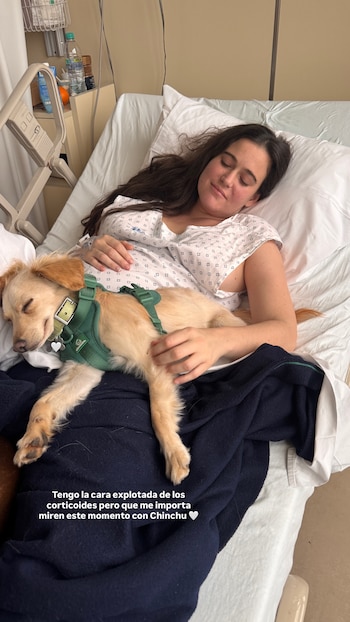 Una mujer con bata hospitalaria yace en una cama clínica con un perro pequeño de pelaje claro y arnés verde sobre su pecho
