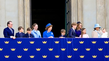 Un grupo grande de adultos y niños de diversas edades está de pie en un balcón detrás de una barandilla azul decorada con coronas doradas