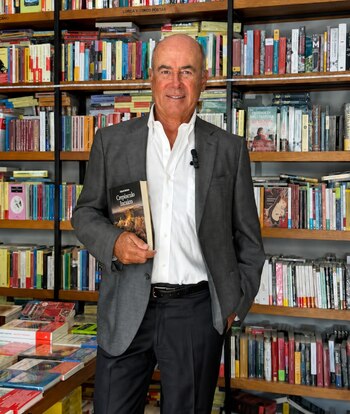 Un hombre de saco gris y camisa blanca sostiene un libro frente a estanterías repletas de libros