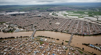 Vista aérea de una ciudad con un río desbordado, barrios residenciales y campos agrícolas inundados, carreteras y puentes cubiertos por el agua, cielo nublado.