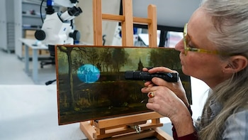 Primer plano de una conservadora con gafas y cabello gris, iluminando una pintura con una linterna que proyecta un círculo azul en el lienzo