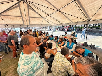 Una gran carpa blanca con armazón metálico cubre varias mesas y personas sentadas y de pie. Varias personas usan chalecos azules. Hay una multitud alrededor