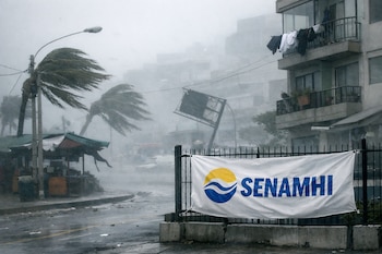 Una pancarta de Senamhi atada a una reja, palmeras y un cartel ladeados por el viento en una calle urbana con edificios, bajo un cielo nublado y con neblina.