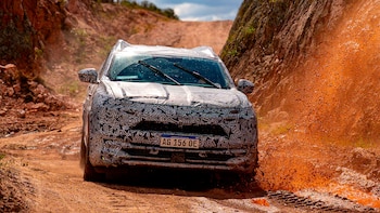 Una camioneta camuflada con matrícula argentina circula por un camino de tierra, salpicando barro en el lado derecho. Hay una pared rocosa rojiza a la derecha