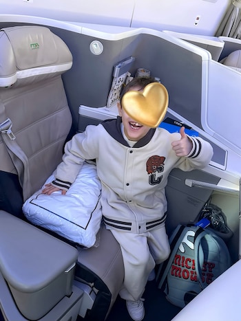 Un niño en un asiento de avión tiene el rostro cubierto por un corazón dorado y realiza un gesto con el pulgar hacia arriba