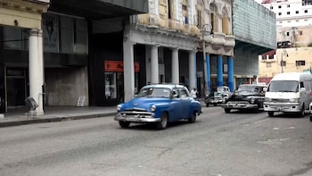 Un coche clásico azul en movimiento por una calle urbana, con otros vehículos y edificios con columnas y balcones al fondo