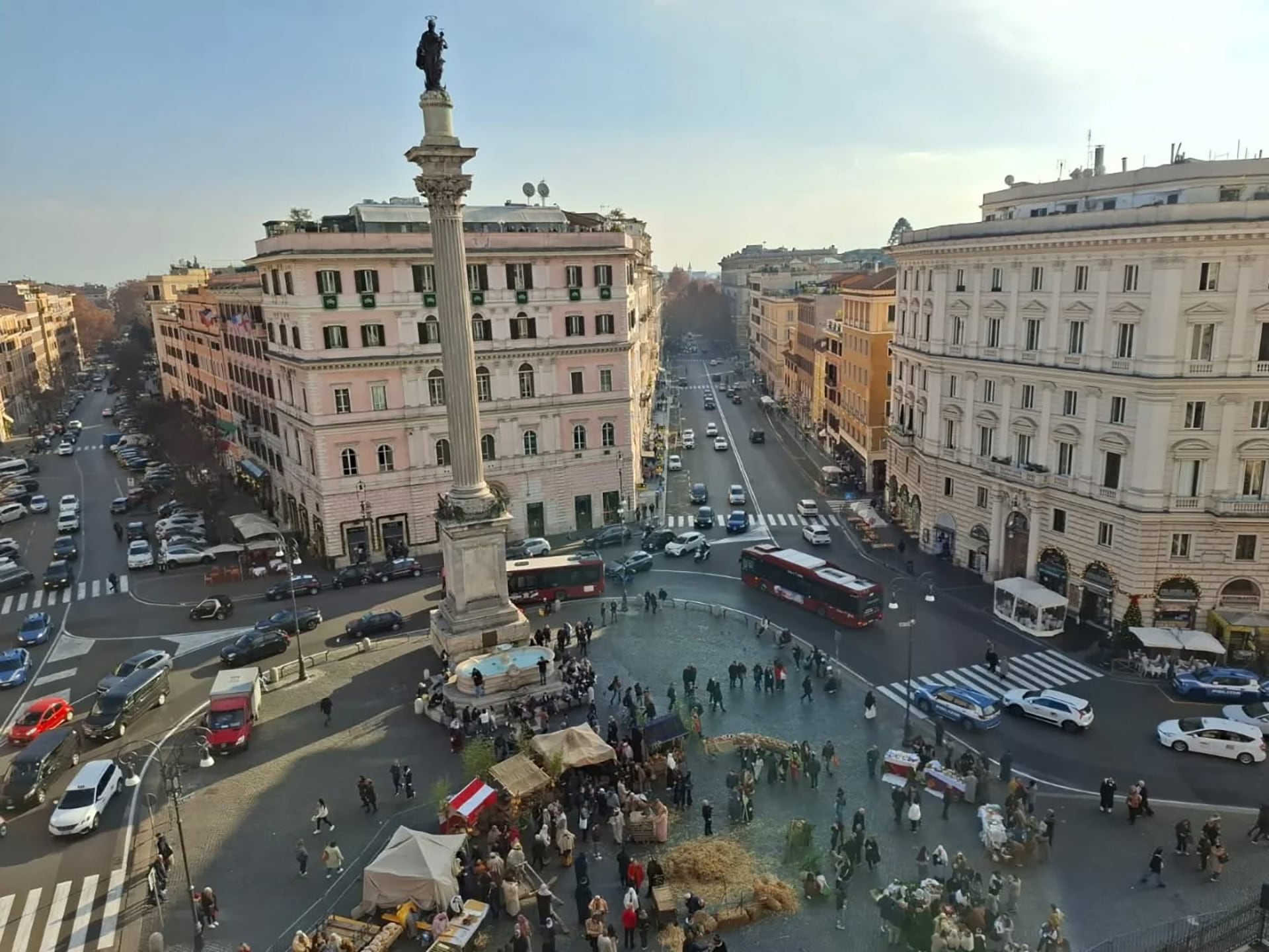 Una vista aérea capta la bulliciosa Plaza Mignanelli en Roma, mostrando la histórica Columna de la Inmaculada Concepción, rodeada de transeúntes, puestos y vehículos en un día soleado.