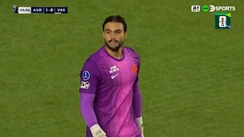 Un portero de fútbol masculino con barba y camiseta morada en un campo verde, con un marcador en pantalla mostrando 'AUD 1-0 VAS'