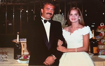 Grecia Colmenares, en vestido blanco, y Marcelo Pelegri, en esmoquin, posan junto a una mesa con champán y aperitivos. Fondo oscuro con luces
