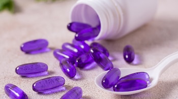 Cápsulas blandas violetas dispersas sobre una mesa junto a un frasco blanco inclinado y dos cápsulas dentro de una cuchara.