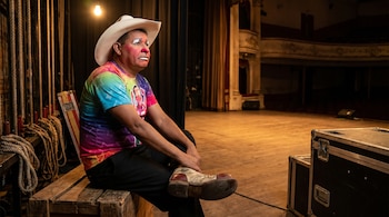 Hombre con maquillaje de payaso y sombrero de vaquero, sentado en un banco de madera tras bambalinas de un teatro, con equipo y cuerdas de escenario al fondo.