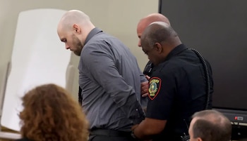 Un hombre calvo con camisa gris es esposado por dos agentes de policía, uno de ellos con uniforme de sheriff de Texas