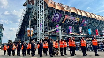 Policías y seguridad con chalecos naranjas y perros K9 saludando frente al Estadio GNP Seguros, con techos dorados, logo GNP y banners de BTS.