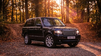 Un vehículo Range Rover SUV negro estacionado en un sendero de tierra cubierto de hojas secas, con árboles de hojas doradas y luz solar brillante al fondo.