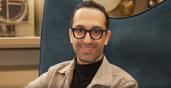 Hombre de tez clara con gafas y barba sonriendo, sentado en un sillón azul. Viste un cuello alto negro y una chaqueta de espiga