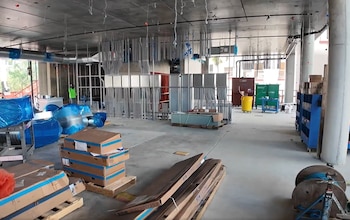 Interior de un edificio en construcción con techos de concreto expuestos, conductos metálicos, tuberías, cajas apiladas, herramientas y un trabajador