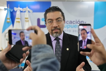 Hombre con barba y gafas, vestido de traje y corbata, hablando. Dos manos con teléfonos móviles en primer plano grabándolo, mostrando su imagen en las pantallas
