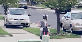 Joven con casco y camiseta gris montando una minimoto roja en un sendero. Al fondo, un coche plateado y uno beige en una calle residencial