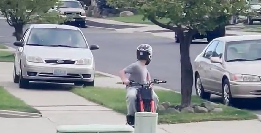 Un joven con casco monta una minimoto roja y negra en un sendero de una calle residencial, con varios coches estacionados o circulando de fondo (Cindy Hagan)