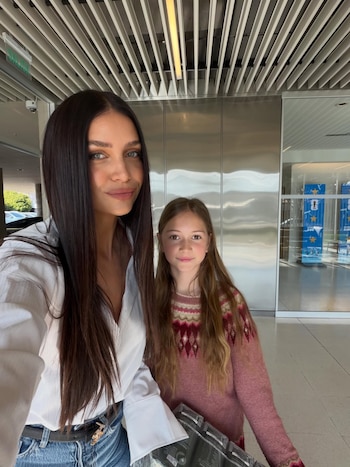 Zaira Nara sonriendo, de cabello oscuro y camisa blanca, junto a una niña con suéter rosa y cabello largo. Fondo interior de edificio moderno
