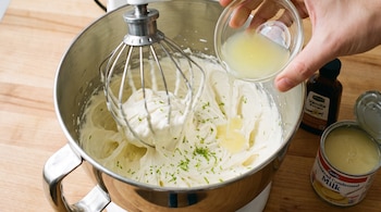 Batidora de pie con batidor de varillas, tazón de acero inoxidable con crema, mano vertiendo jugo de limón, ralladura verde, esencia de vainilla, leche condensada.