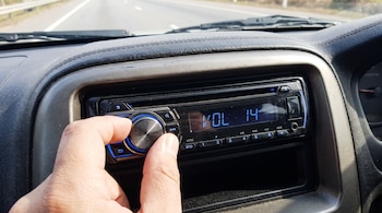 Una mano ajusta una perilla plateada de un estéreo de automóvil. La pantalla digital muestra "VOL 14". Se ven parte del tablero y una carretera al fondo.